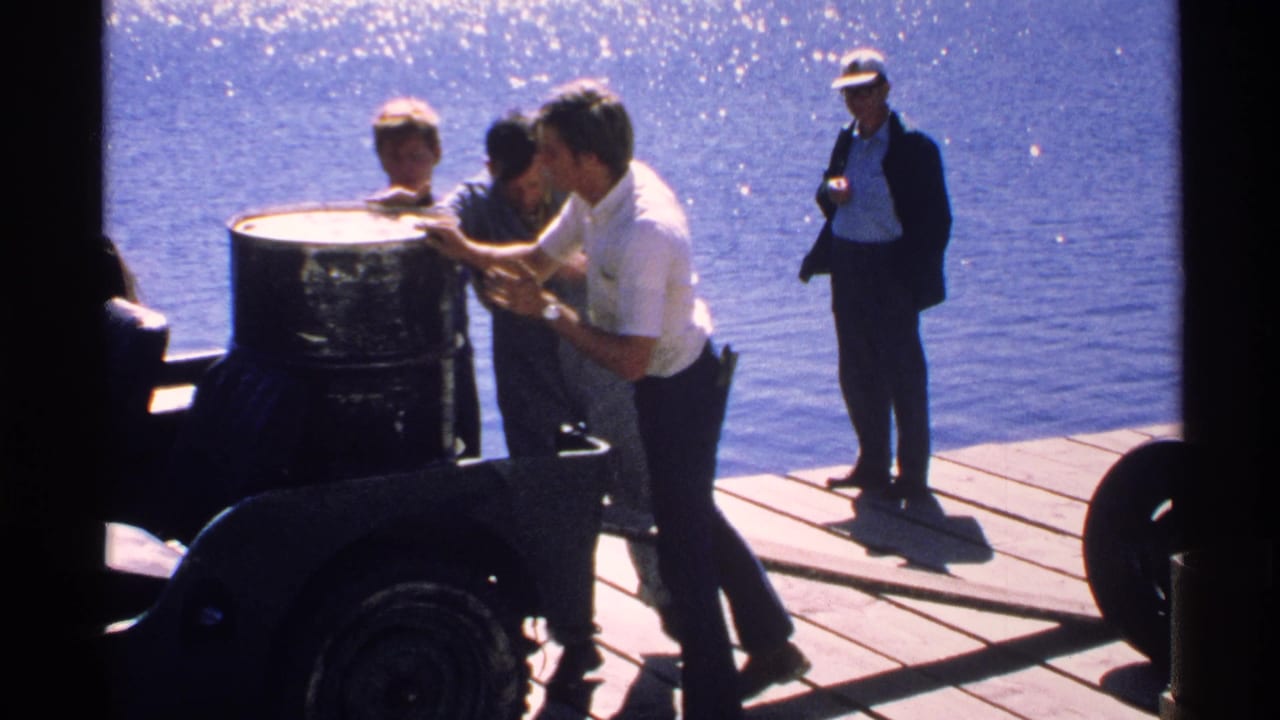 Passengers stand on a wooden boat deck near a large barrel beside bright blue water.