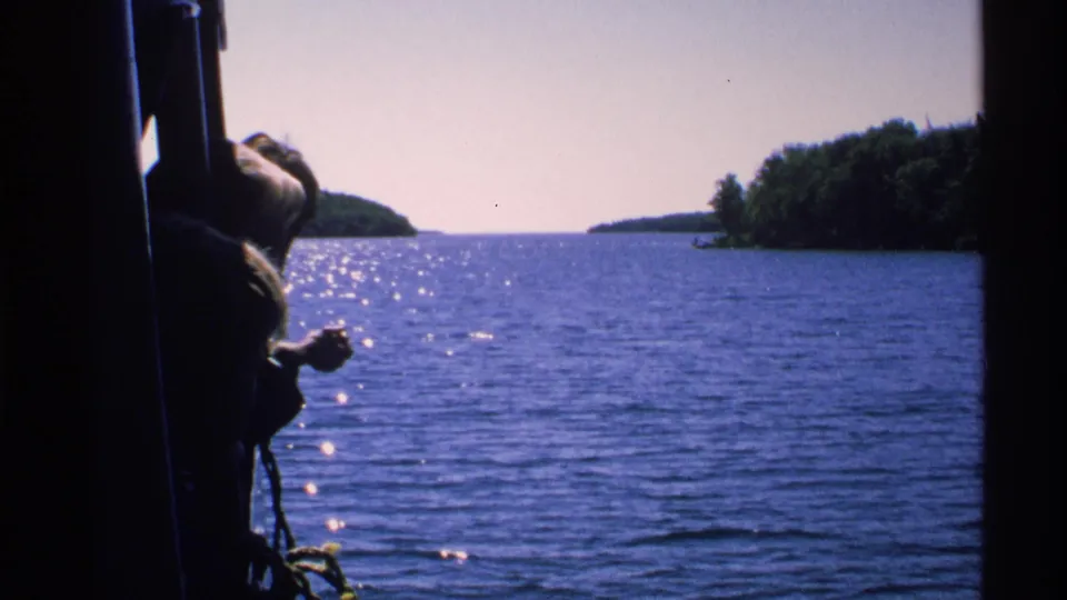 Handheld 8mm view from a boat at Isle Royale, with a passenger partly silhouetted beside sunlit water and wooded shoreline.