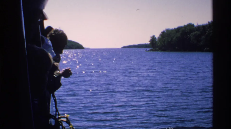 Boat-side home-movie view of a passenger near the frame edge with Isle Royale water and forested islands in the background.