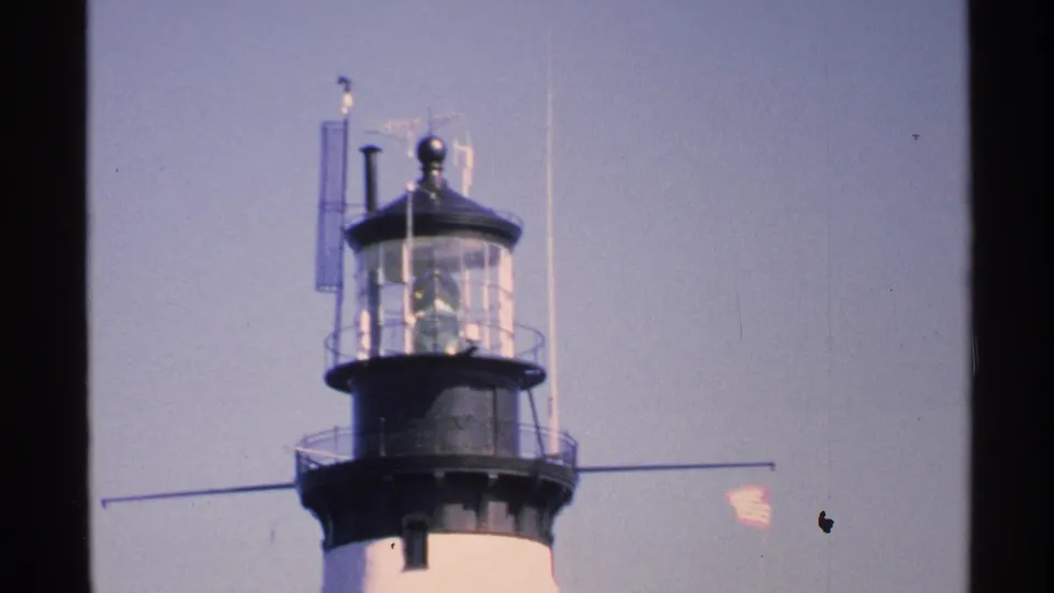 1972 Isle Royale 8mm frame showing a lighthouse lantern room, railings, small flag, and pale sky.