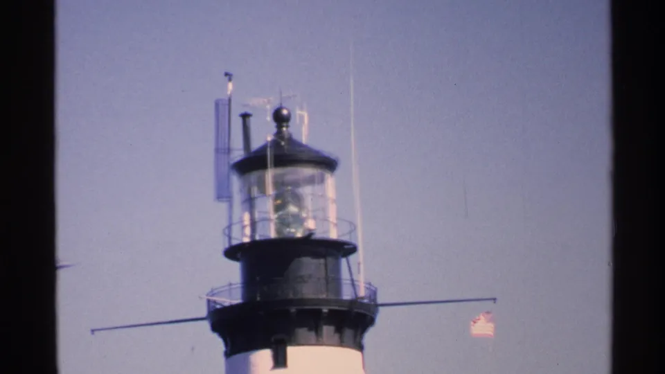 1972 Isle Royale home-movie frame of a lighthouse lantern room with railings, rods, a small flag, and open sky.