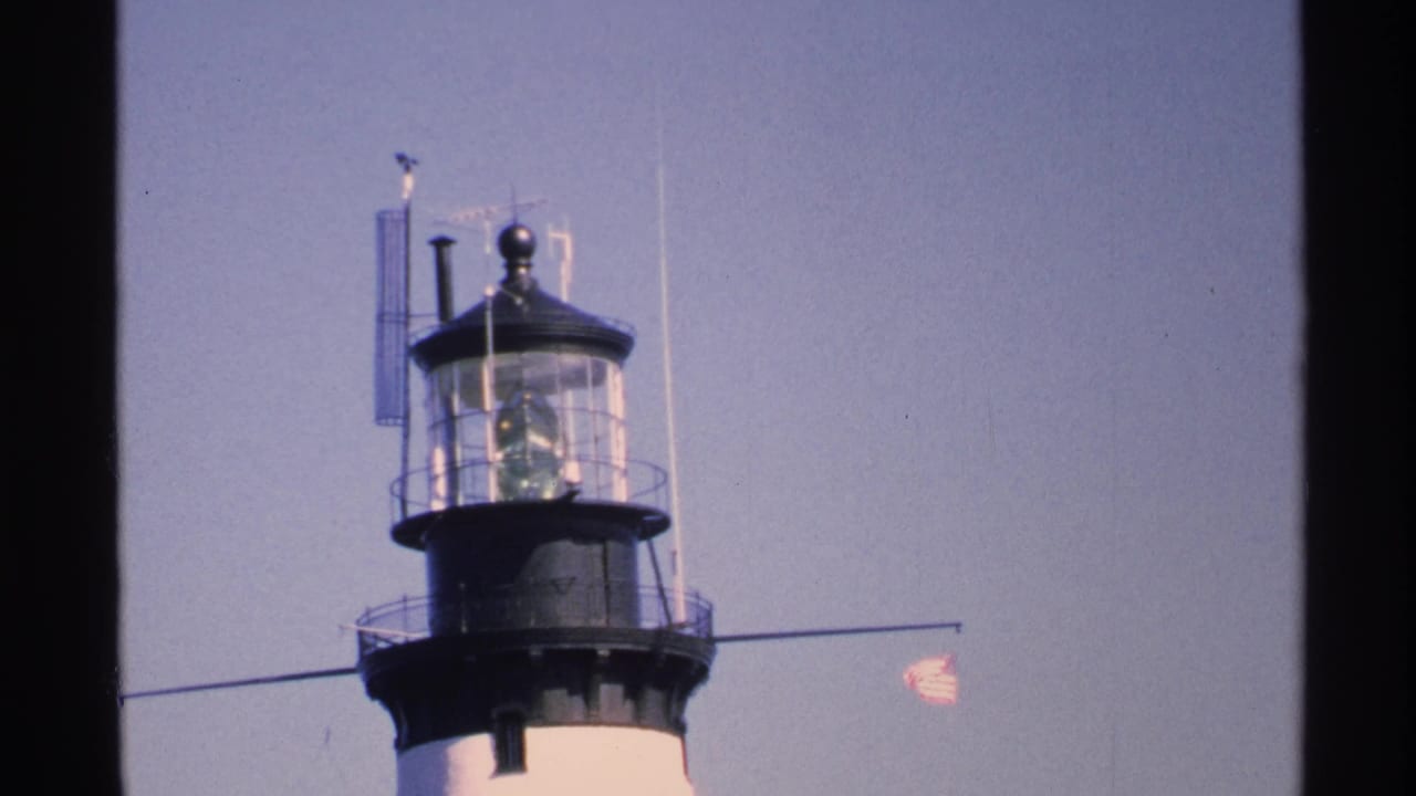 Close view of a lighthouse lantern room and small American flag against pale blue sky.