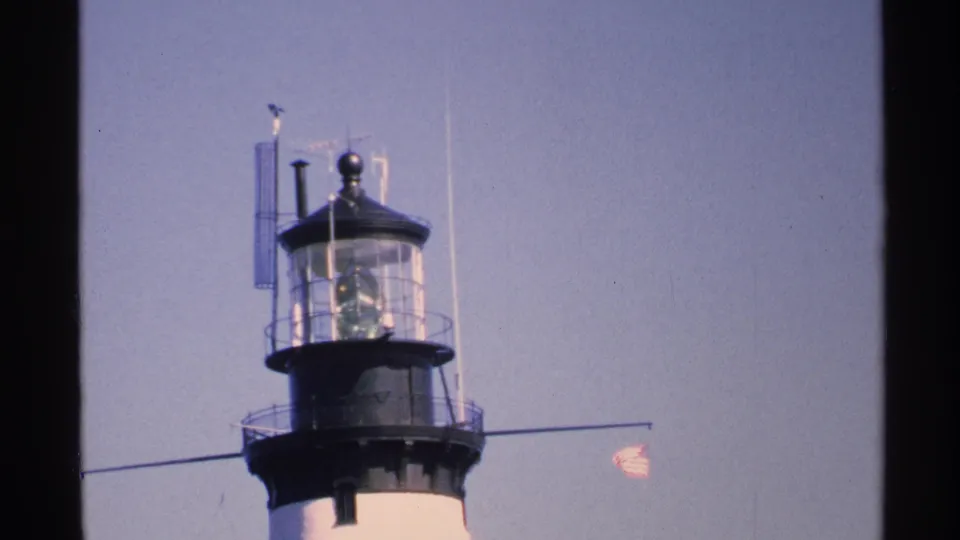 1972 Isle Royale home-movie frame showing the upper lighthouse tower, black lantern room, railings, and small American flag.