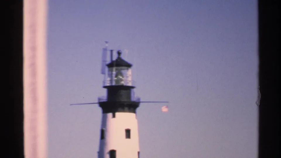 1972 Isle Royale frame showing the lighthouse tower and lantern room with a small flag, softened by 8mm film grain.