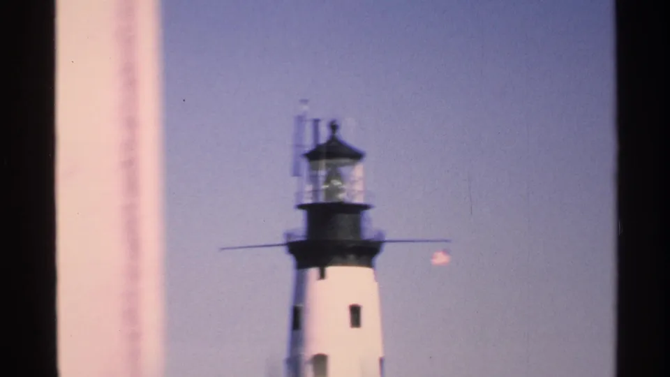 1972 Isle Royale home-movie frame showing a white lighthouse top with a black lantern room against a blue sky, partially framed by dark edges.