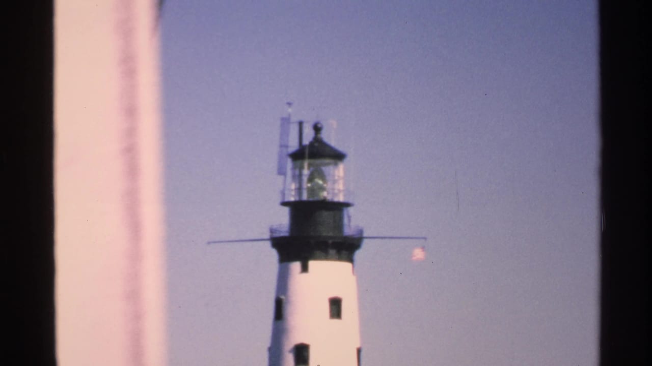 1972 Isle Royale footage frame with the lighthouse upper tower centered between dark vertical edges and a pale blue sky.