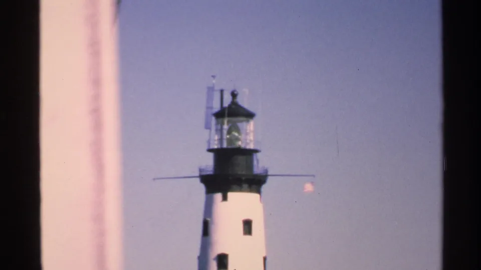 1972 Isle Royale footage frame with the lighthouse upper tower centered between dark vertical edges and a pale blue sky.