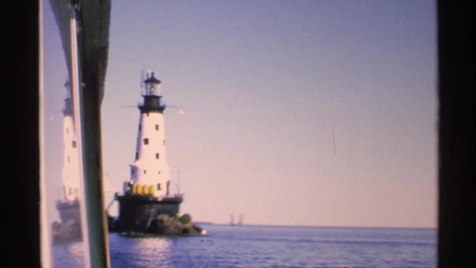 1972 Isle Royale home-movie frame showing the lighthouse on rocks in blue water, with window or boat structure along the left edge.