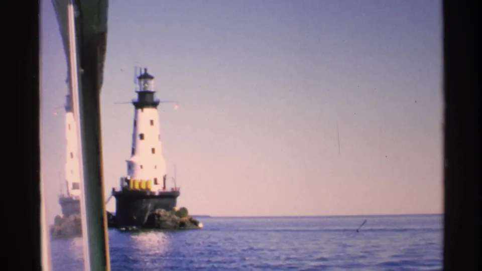 1972 Isle Royale frame with the lighthouse set back on a rocky base, open water in front, and a wide blue sky beyond.