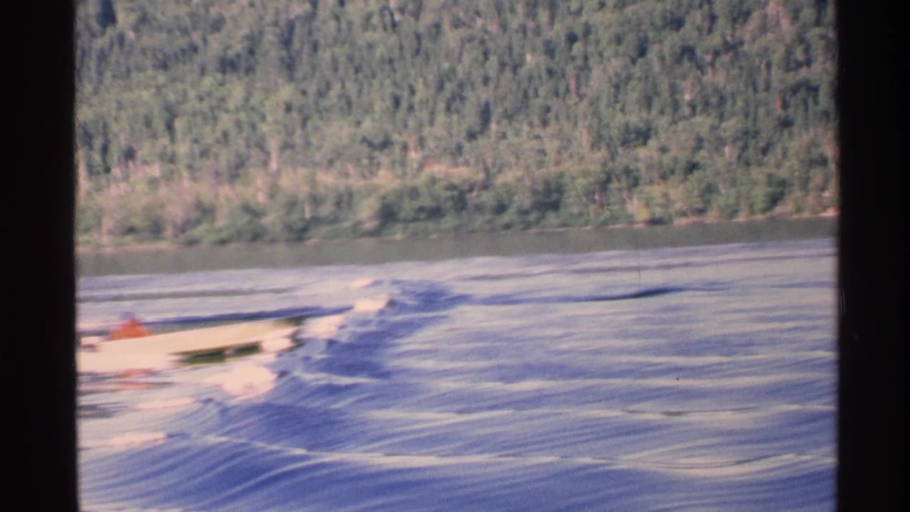 Small motorboat crosses a lake near a forested shoreline with dark film borders.