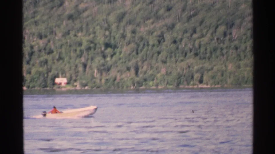 A small boat with a distant seated person travels along calm Isle Royale water, with forest and a red structure beyond.