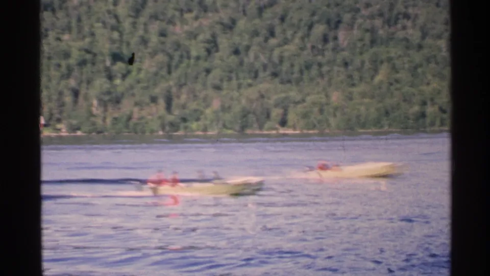 1972 Isle Royale home-movie frame showing two yellow speed boats crossing a calm lake before a wooded shoreline.