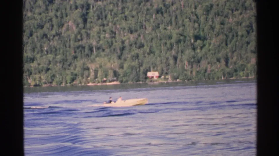 The yellow boat appears farther from the camera on a calm lake, with passengers, low wake, forested hills, and a small lakeside building visible.
