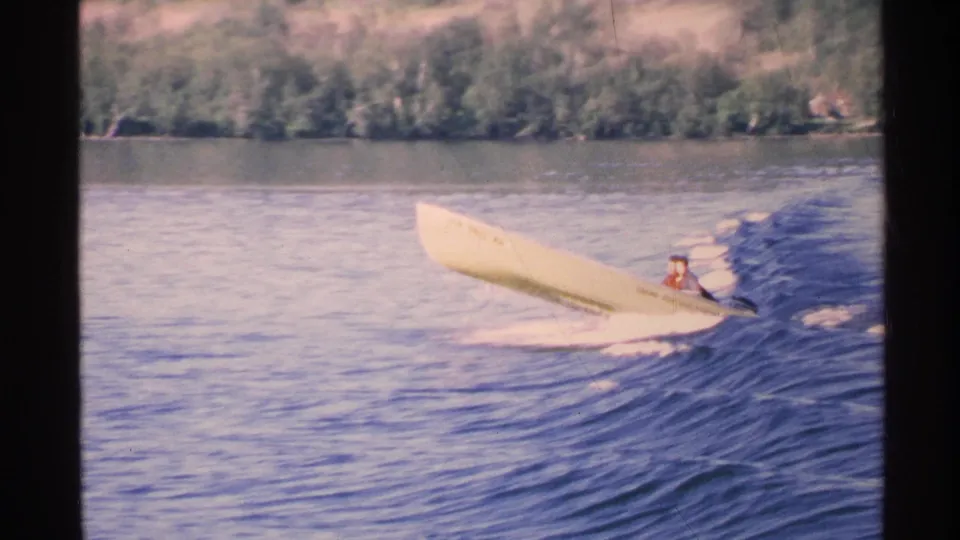 Close 1972 8mm view of a yellow speed boat banking over a wake on Isle Royale water, with a passenger visible and trees beyond.