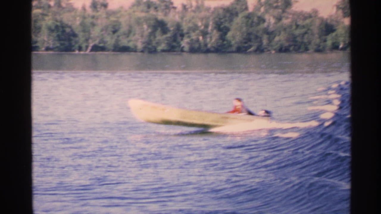 Small motorboat crosses a lake beside a high white wake and wooded shoreline.