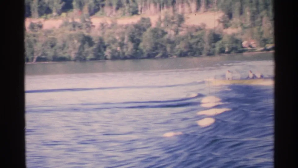 1972 Isle Royale home-movie frame with a distant motorboat near the right edge, a trailing wake, and a wooded shoreline beyond.