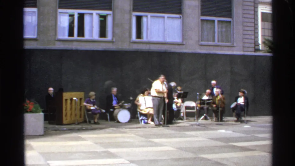 A small outdoor band performs before a building, with seated musicians, a drum set, and a man standing at a microphone.
