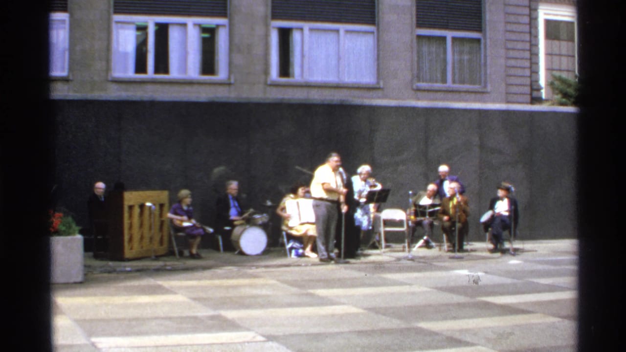 Outdoor ensemble with vocalist, drummer, and seated musicians in front of windows.