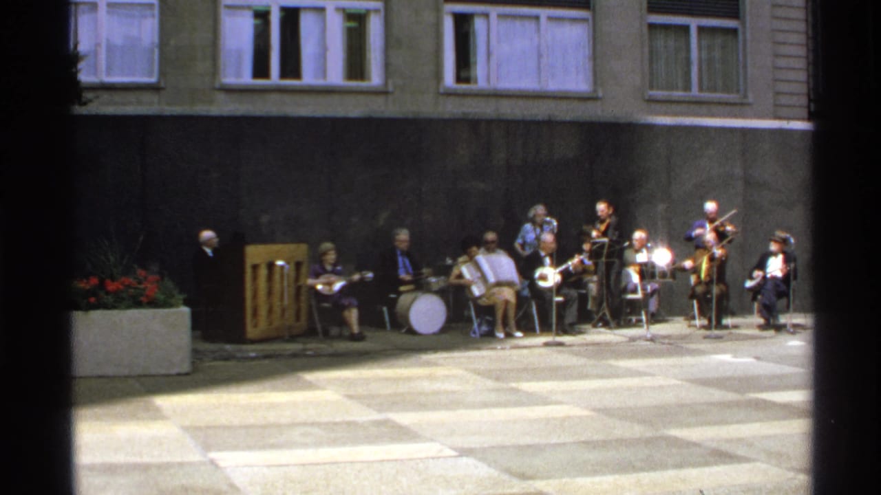 Small outdoor band seated in a checkered courtyard with piano, drum, accordion, and string instruments.