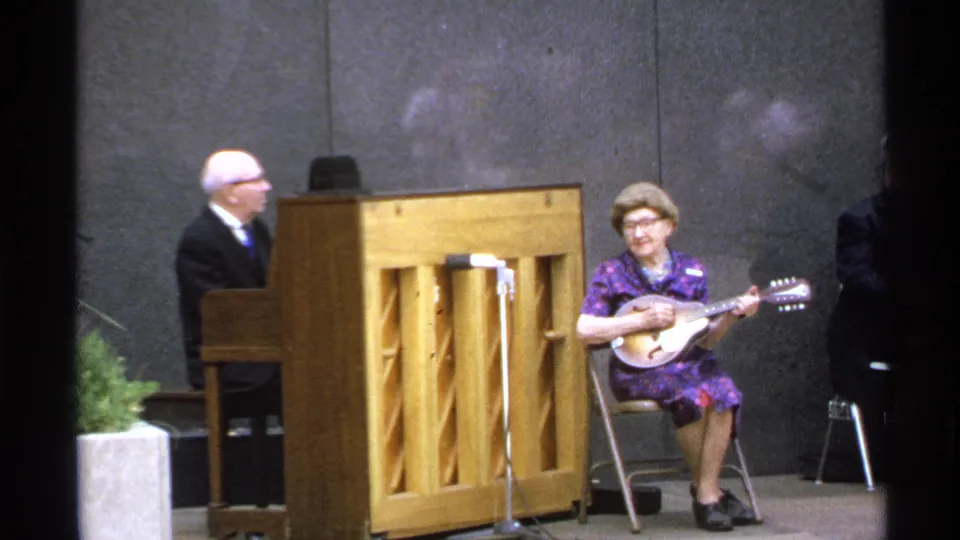 Close 1972 Isle Royale frame of a woman in a purple dress playing mandolin with piano accompaniment outdoors.