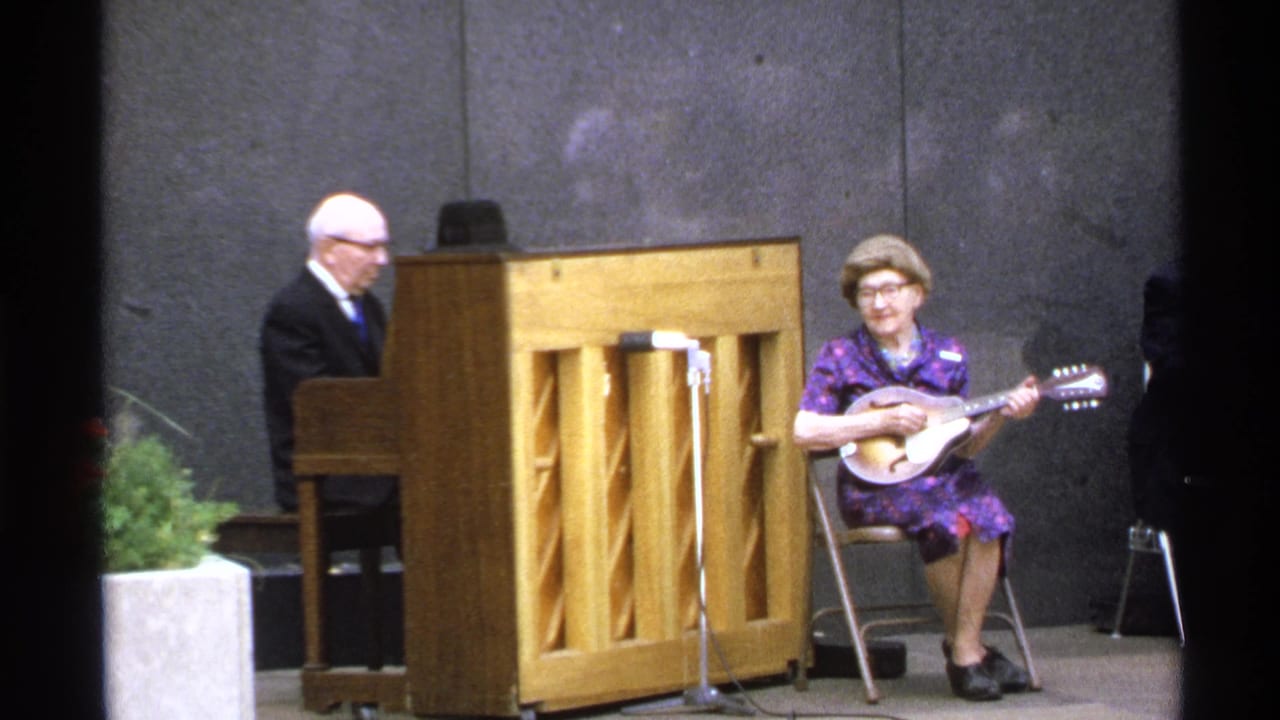 Man at upright piano beside seated woman playing mandolin on a small stage.