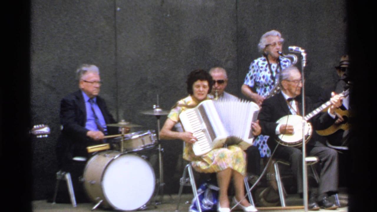 1972 Isle Royale 8mm frame showing a folk ensemble performing on a simple stage with accordion, banjo, drums, strings, and microphones.