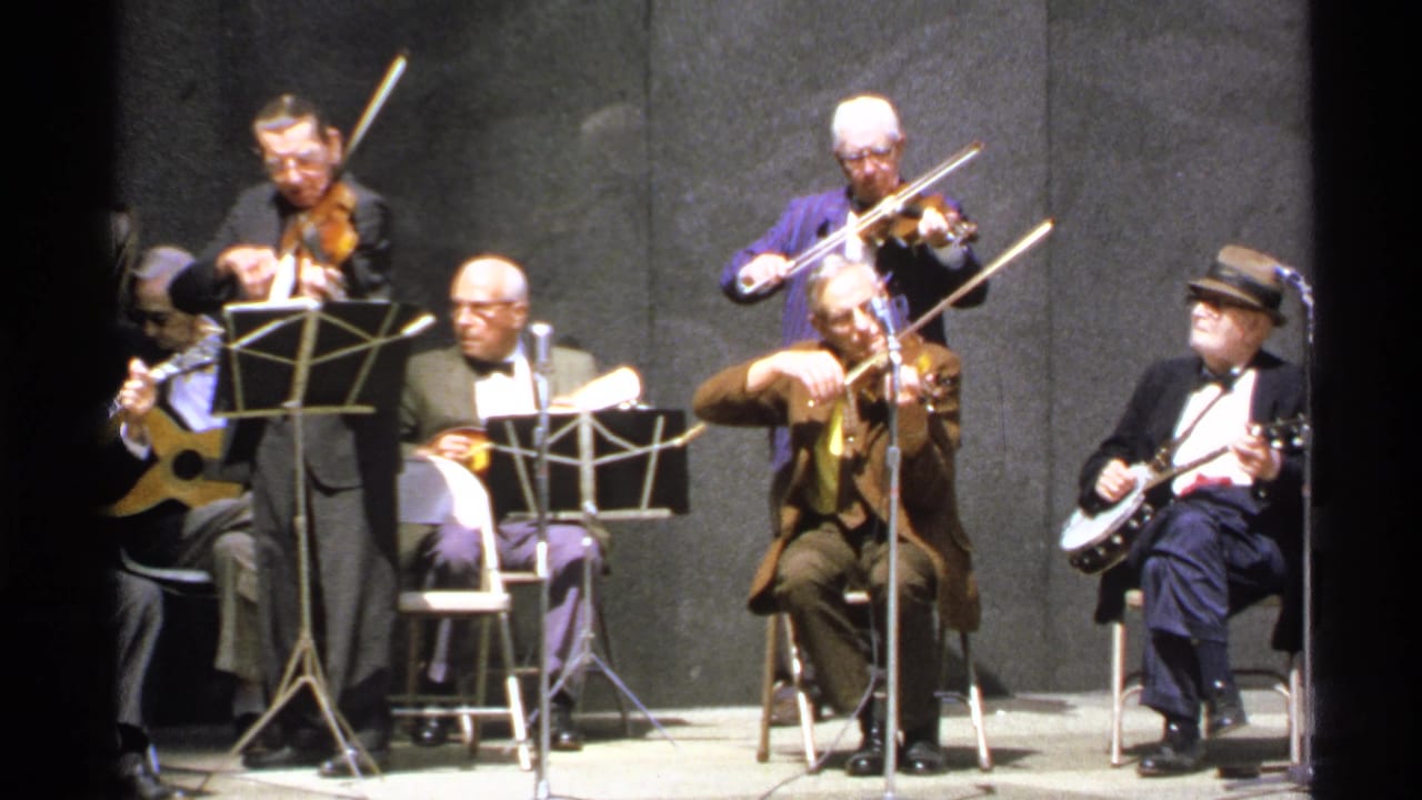 Violinists, seated string players, music stands, microphones, and a banjo player on stage.
