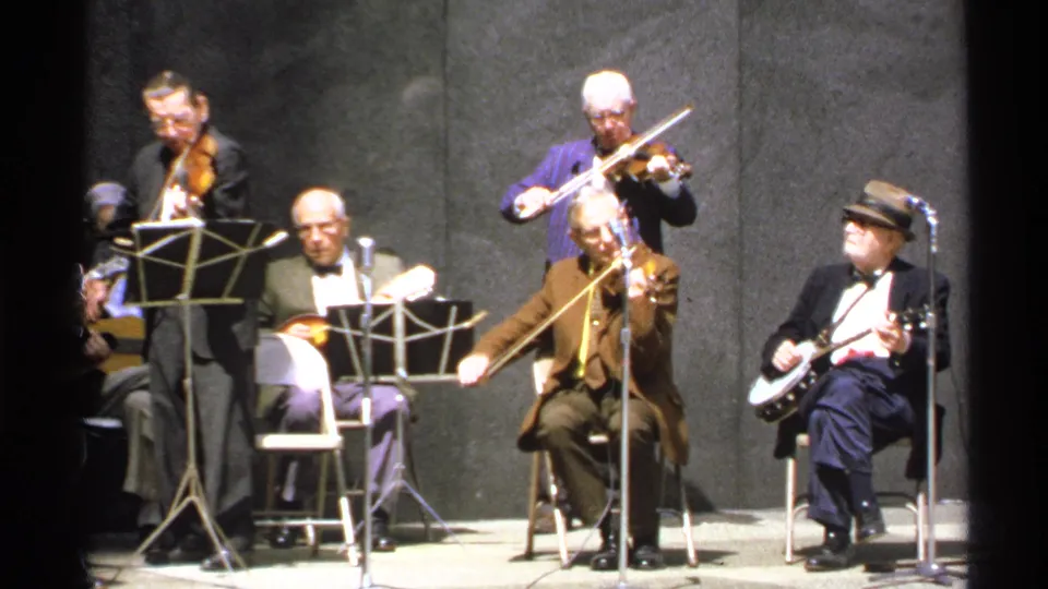 1972 Isle Royale 8mm frame focused on violinists and seated folk musicians performing beside music stands and microphones.
