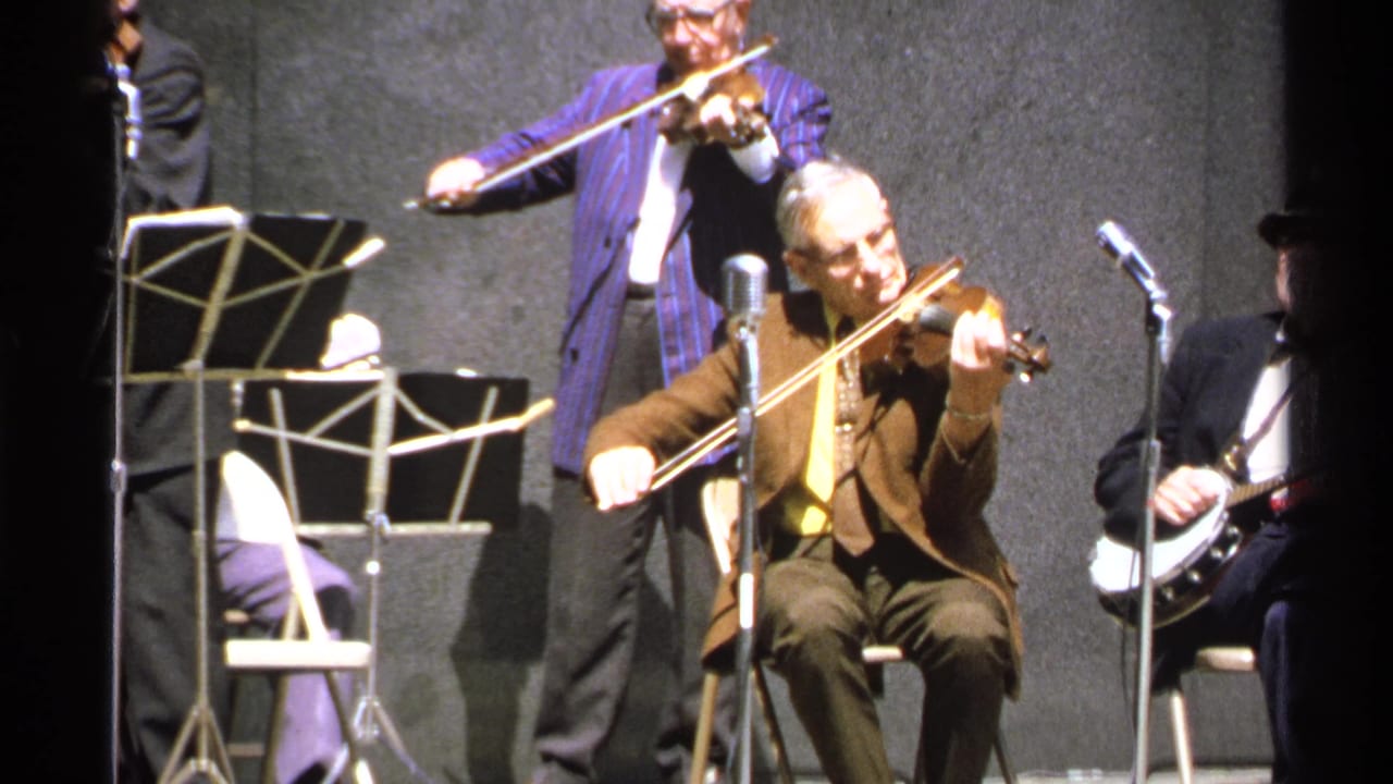 A seated violinist plays beside microphones while other musicians stand nearby.