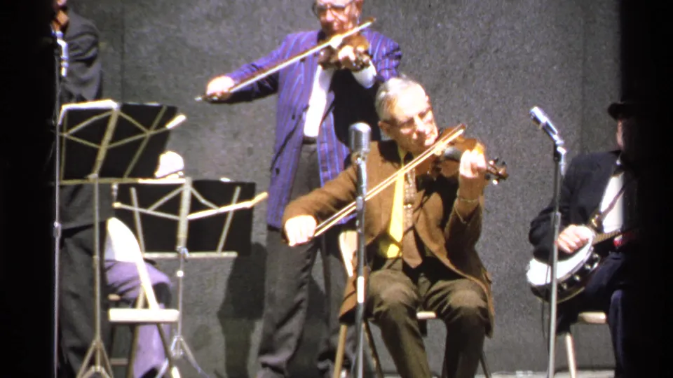 A seated violinist in a brown suit plays on stage as another violinist and a banjo player appear near microphones.