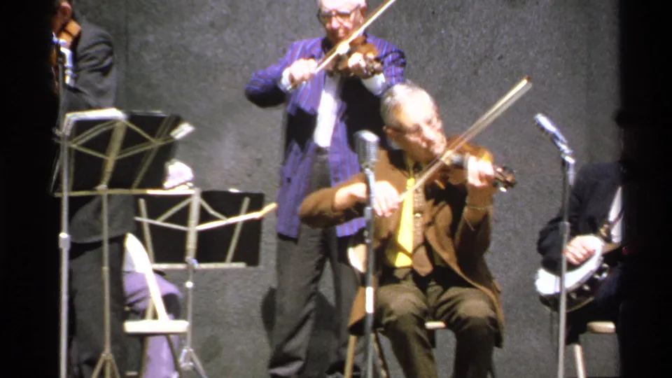 The 8mm frame moves closer to the violinists, with music stands, microphones, and part of a banjo player visible at the edge.