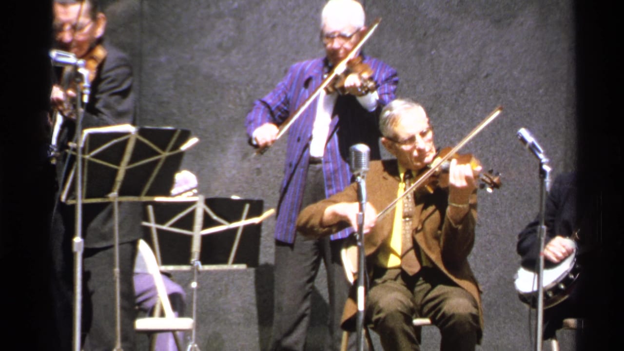 Older men perform on stage with violins, banjo, microphones, and music stands.