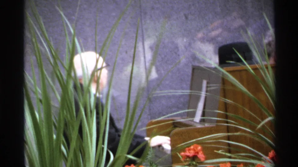 1972 Isle Royale performance frame with tall foreground plants, a partially visible pianist, wooden piano, and dim stage backdrop.
