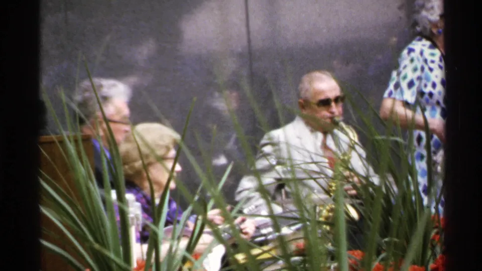 1972 Isle Royale frame showing a saxophonist in a light suit and sunglasses among seated adults at an outdoor performance.
