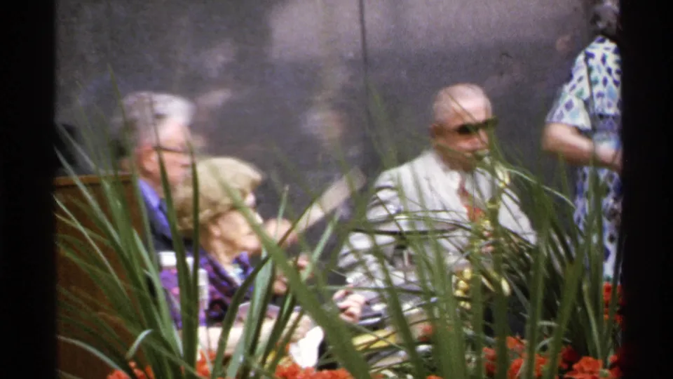 1972 Isle Royale 8mm frame with saxophonist, drummer, seated adults, and planted foreground during a small outdoor music set.