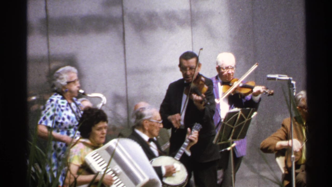 Older adult musicians perform outdoors with accordion, banjo, saxophone and violins near microphones.
