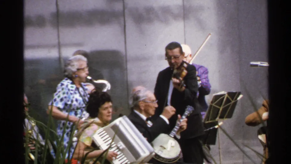 1972 Isle Royale home-movie frame of a small outdoor ensemble playing saxophone, banjo, accordion and violin.