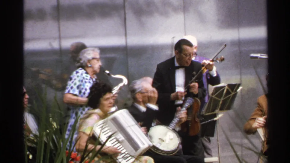 1972 Isle Royale 8mm frame with accordion, banjo, saxophone and violin players performing in a park-like setting.