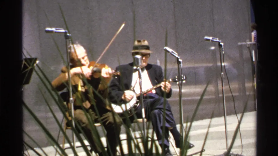 1972 Isle Royale home-movie frame showing a seated banjo player and a violinist performing beside stage microphones.