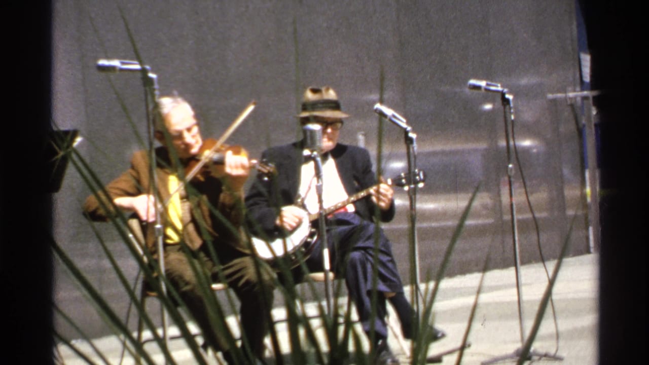 1972 8mm frame of two seated folk musicians, one on violin and one on banjo, performing into stand microphones.
