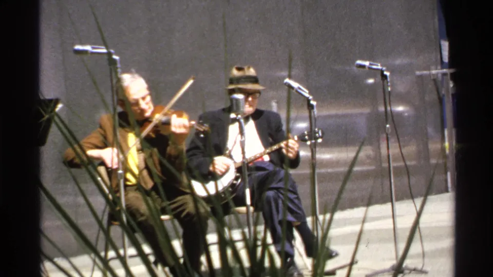 1972 8mm frame of two seated folk musicians, one on violin and one on banjo, performing into stand microphones.