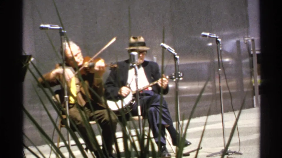 The violinist leans into the instrument while a banjo player in a fedora sits beside vintage-style microphones.