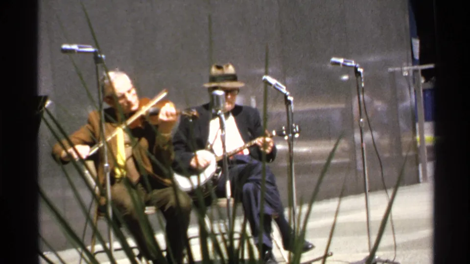 Close 8mm view of two seated folk musicians performing, with tall grass crossing the foreground and microphone stands around them.