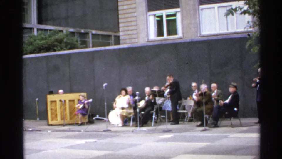 Wider 1972 home-movie frame showing a small acoustic ensemble seated outside with microphones, a piano, and building windows behind them.