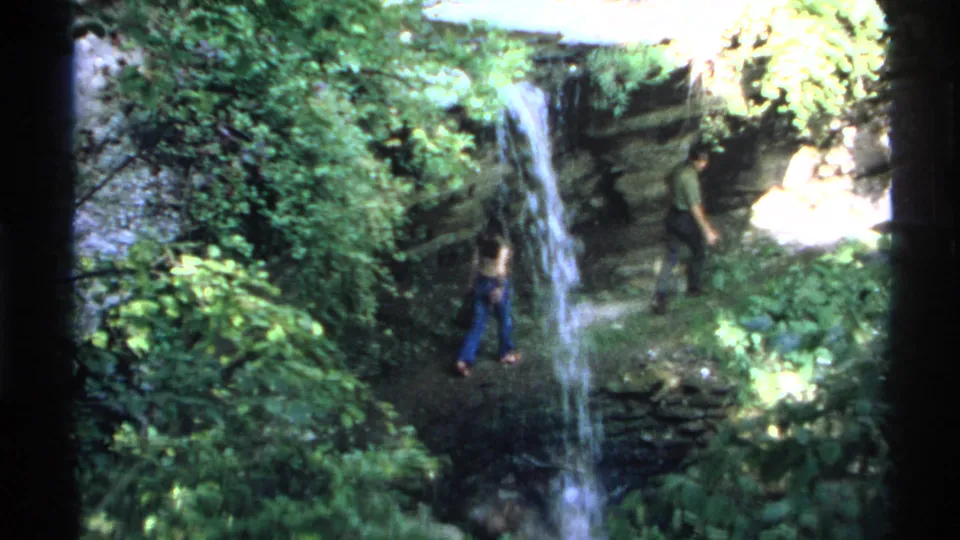 1972 Isle Royale home-movie frame showing a woman and man near a narrow waterfall on a rocky forest path.