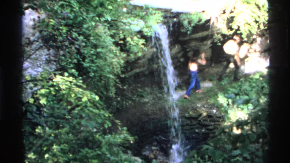 1972 Isle Royale frame with two adults moving through a wooded waterfall area in casual hiking clothes.