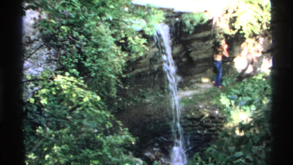 1972 Isle Royale 8mm frame of a narrow waterfall framed by foliage, with a distant visitor standing near the rocky ledge.