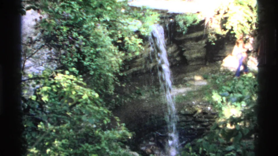1972 Isle Royale home-movie frame showing the waterfall and rocky ledge, with a partially visible visitor near the right edge.
