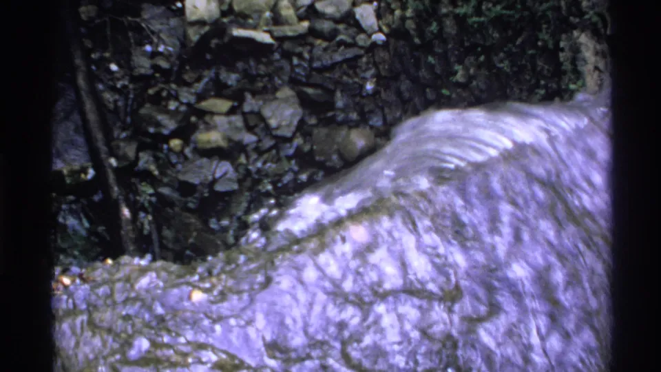1972 Isle Royale 8mm frame showing rushing creek water over rocks near a dark stone embankment and vegetation.