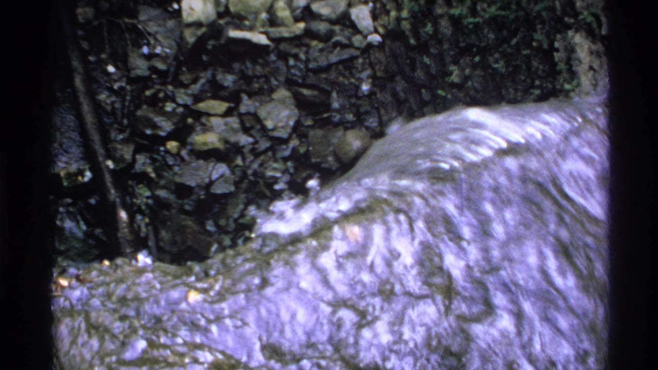 Water rushes across rocks beneath a rough stone bank and leafy growth.
